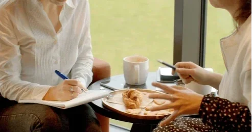 Two women having a conversation at a table with a pastry and cups, one woman is taking notes, and the other is gesturing.