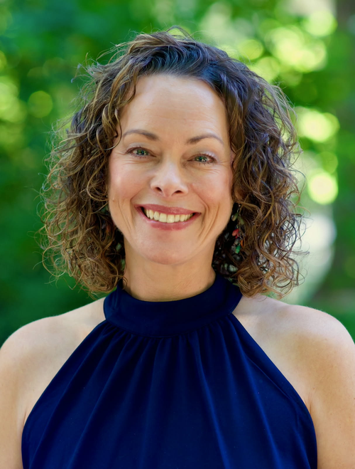 A smiling woman with curly brown hair and light skin, wearing a navy blue sleeveless top, outdoors with green trees in the background.