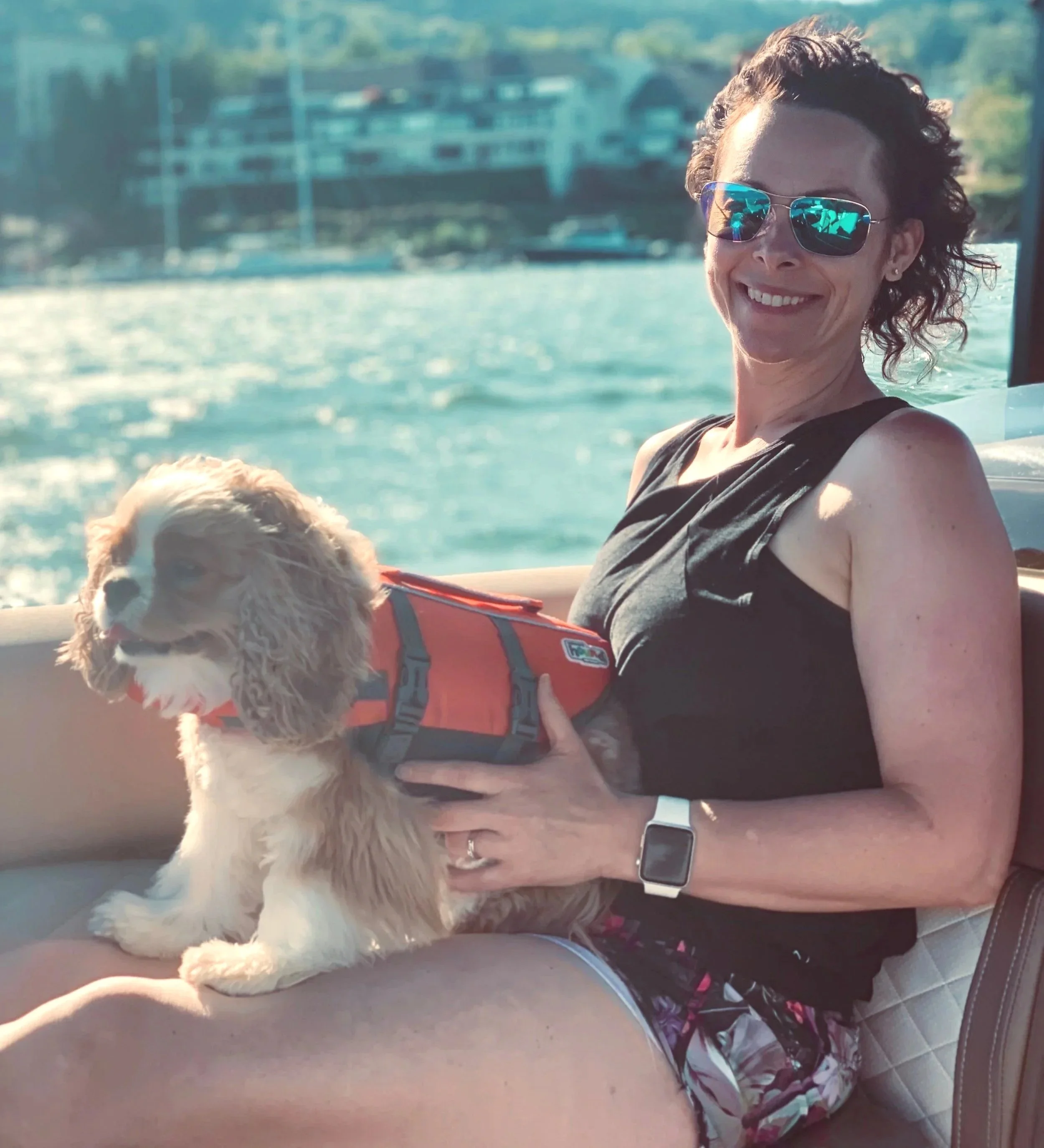 A woman with curly hair wearing sunglasses smiling while holding a dog on a boat, with water and buildings in the background.
