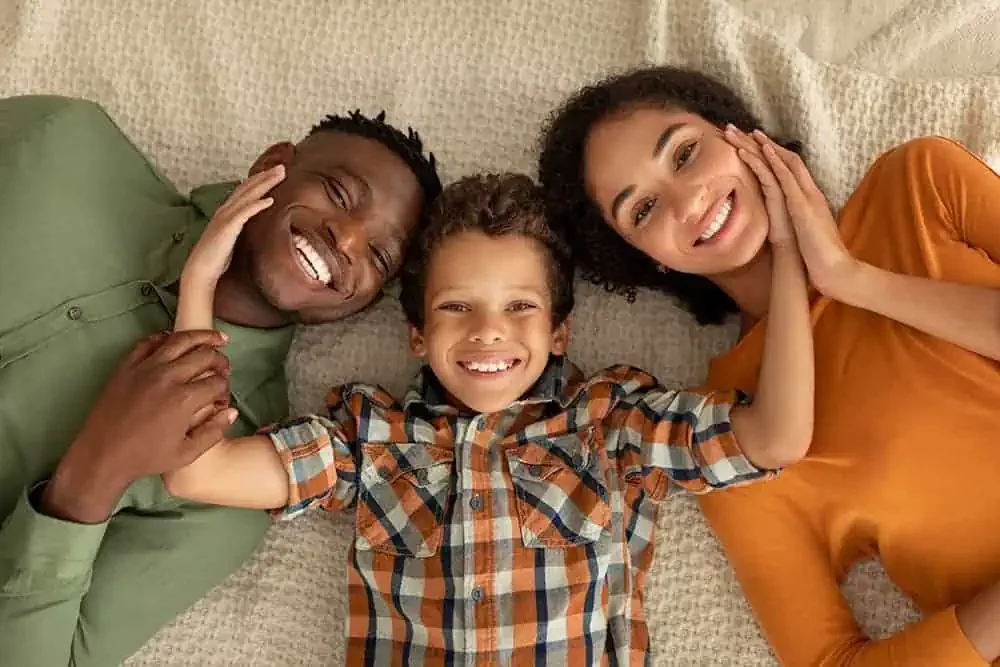A smiling family of three, with a man, woman, and young boy, lying on the carpeted floor with their heads touching, looking up at the camera.