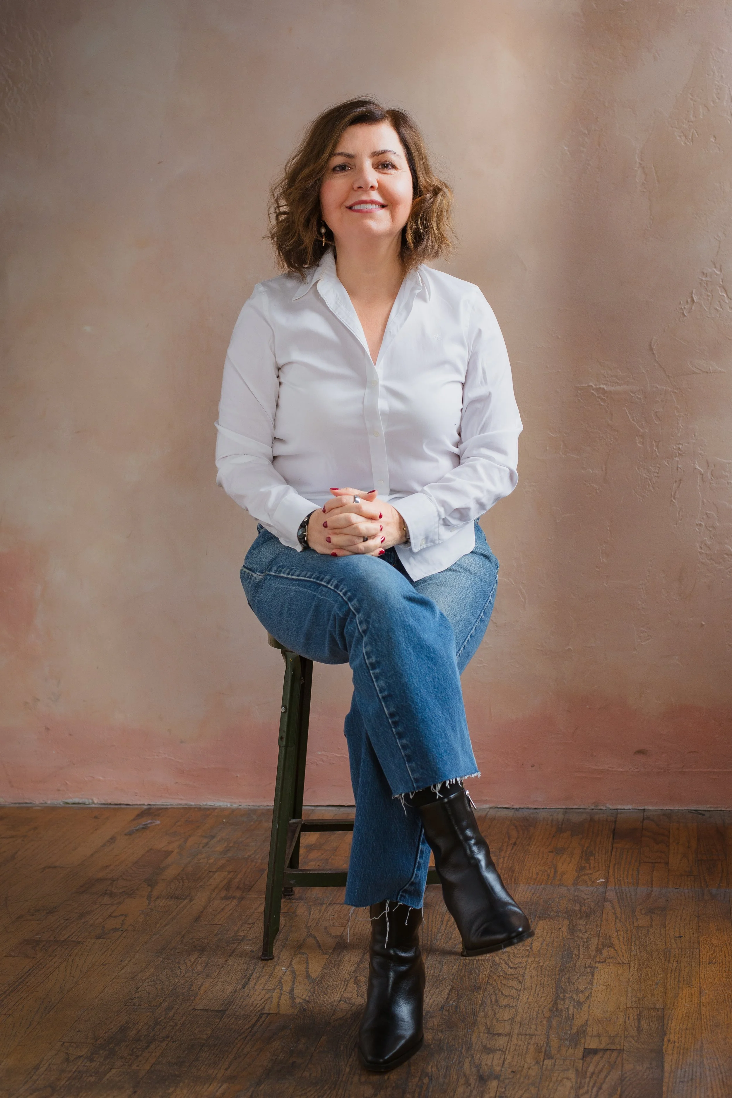 A woman with shoulder-length brown curly hair sitting on a stool against a textured beige wall. She is smiling, wearing a white button-up shirt, blue jeans, and black leather boots. She has her hands clasped in her lap, and her left leg crossed over her right.