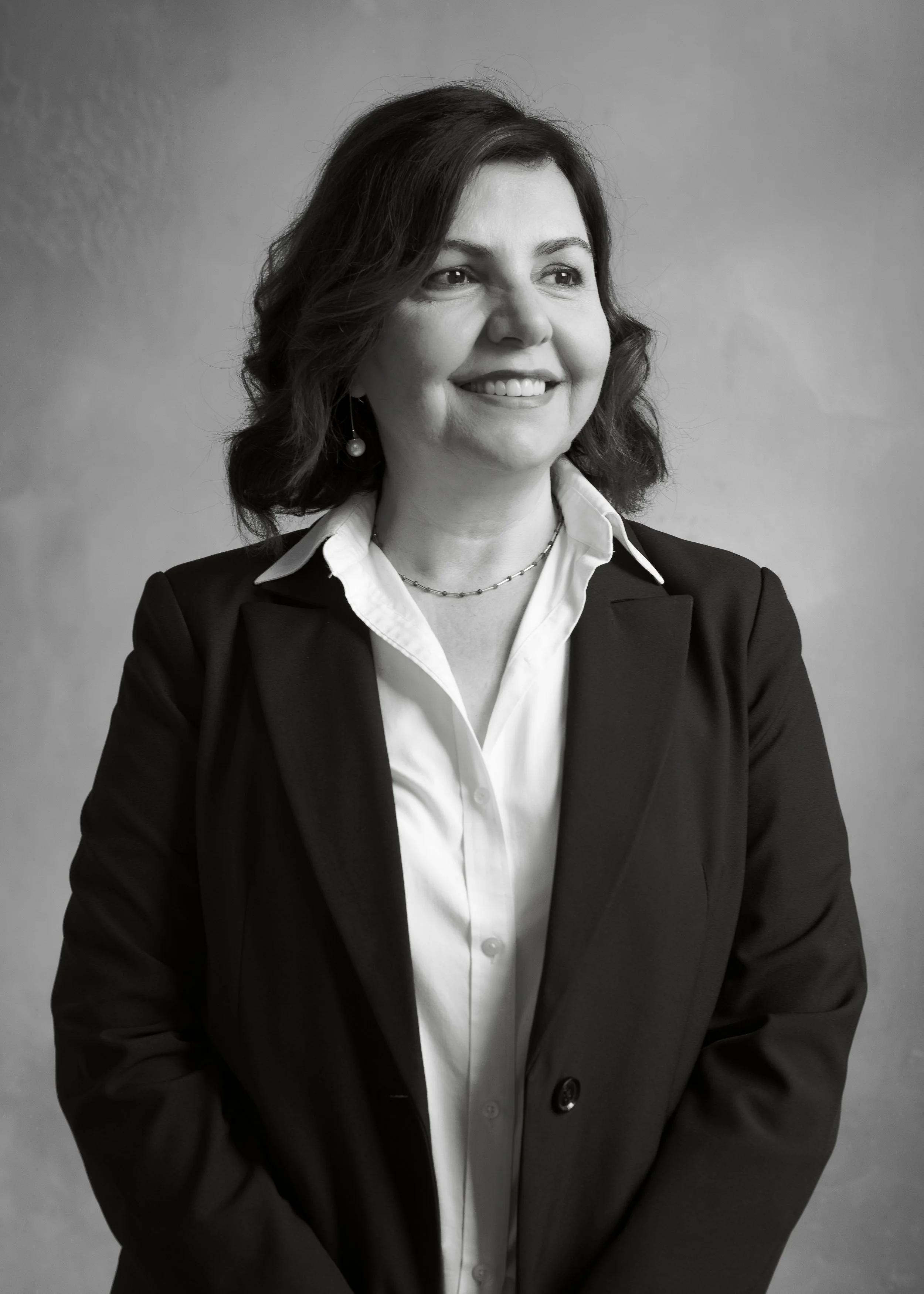 Black and white portrait of a woman with shoulder-length dark hair, smiling, wearing a dark blazer over a white collared shirt with a pearl necklace and earrings, against a plain background.