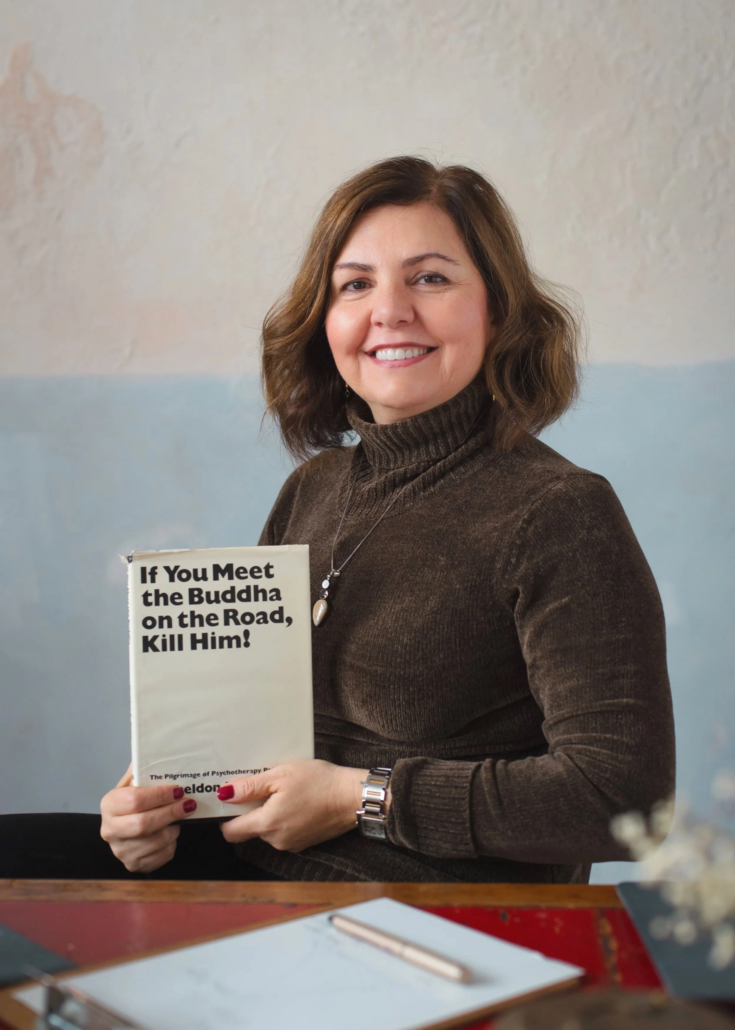 A woman with shoulder-length brown hair smiling, wearing a brown turtleneck sweater, holding a book titled 'If You Meet the Buddha on the Road, Kill Him!' in front of a light-colored wall.