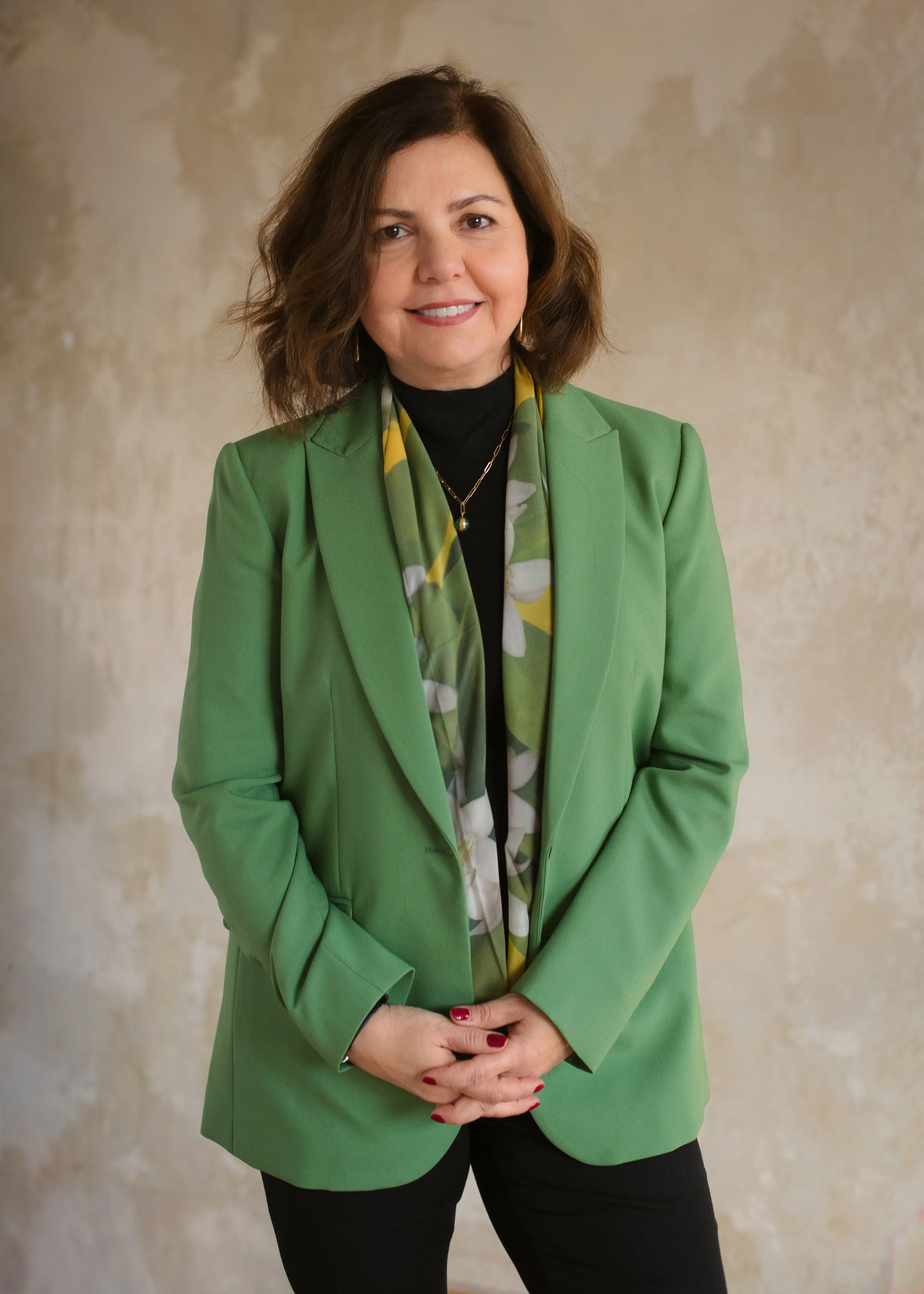A woman with shoulder-length wavy brown hair wearing a bright green blazer, a black turtleneck, layered gold necklaces, and floral scarf, standing against a beige textured wall.