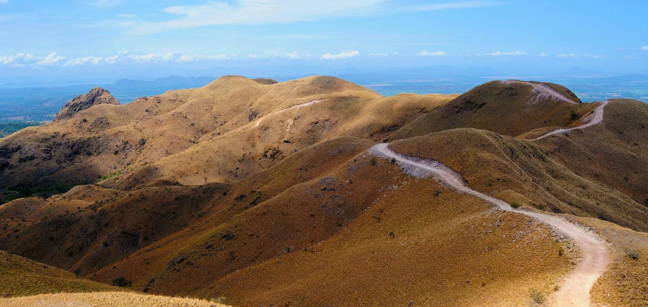 Scenic view of rolling brown hills with a winding dirt trail, under a blue sky with clouds.