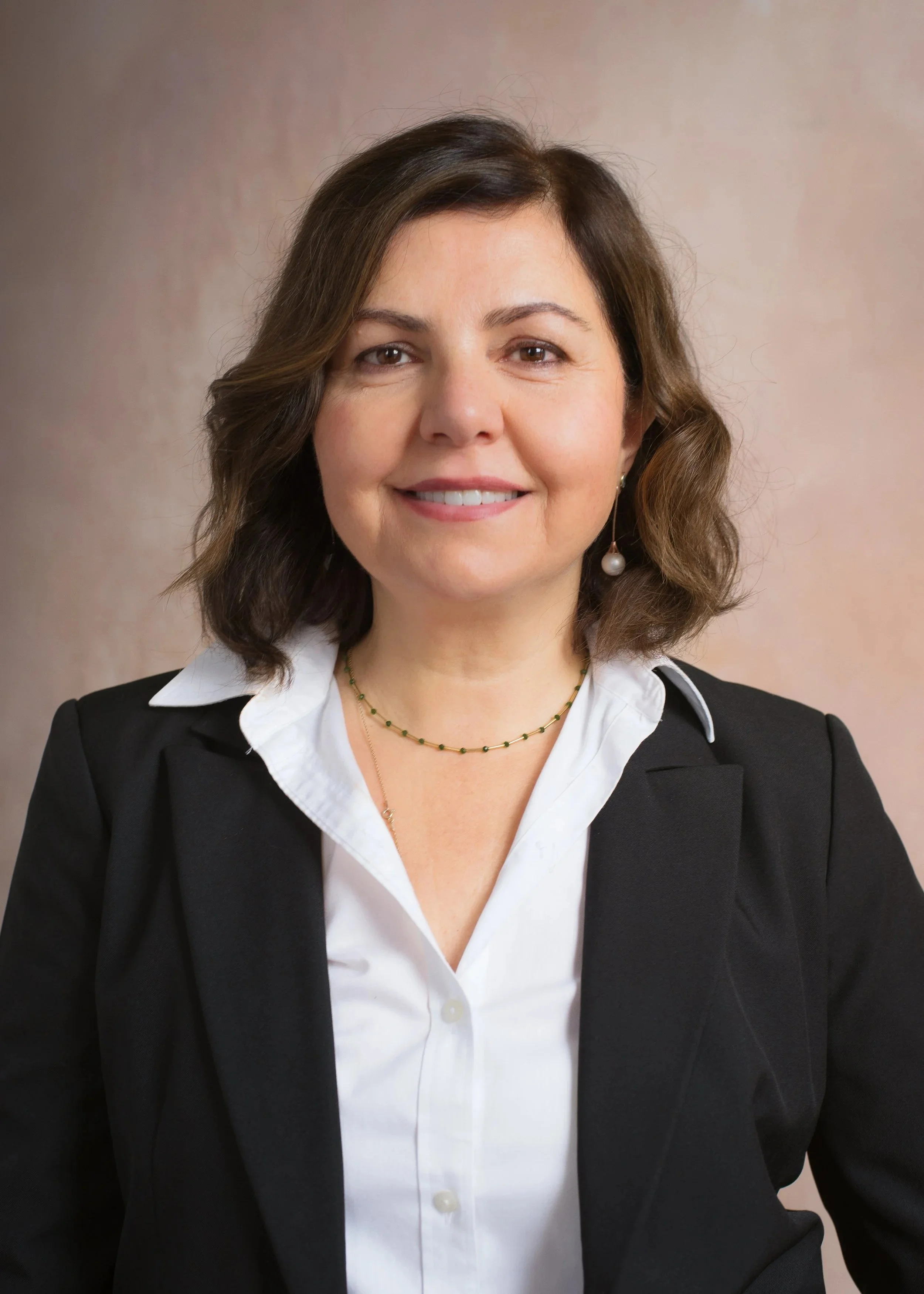 A woman with shoulder-length brown hair, wearing a white blouse, black blazer, gold necklace with small green beads, and pearl earrings. She is smiling and looking at the camera with a neutral background.