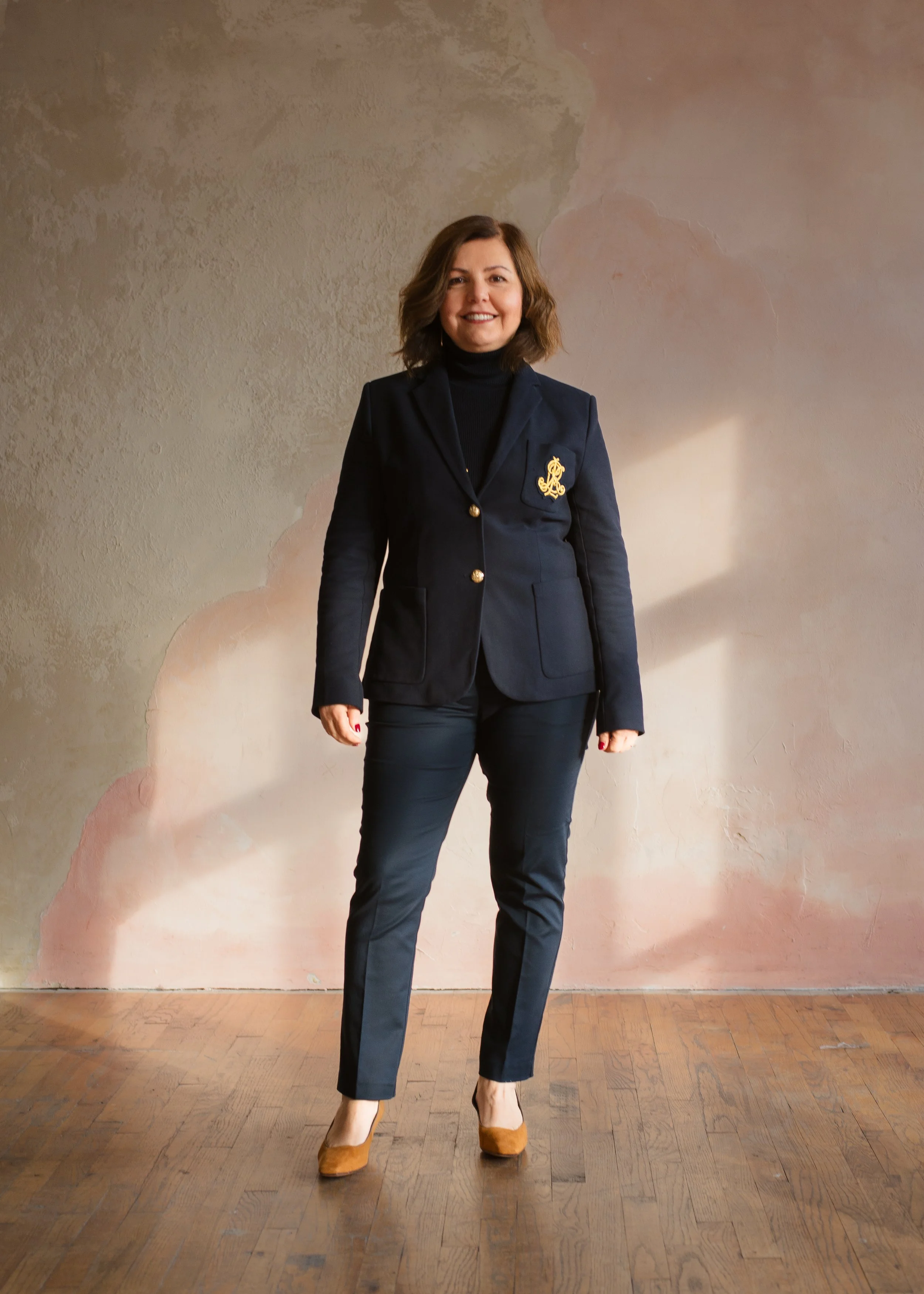 A woman with shoulder-length brown hair smiling, standing on a wooden floor in front of a textured wall with peeling paint. She is wearing a black blazer with gold buttons, black pants, tan high heels, and a black turtleneck, with a gold emblem on her blazer.