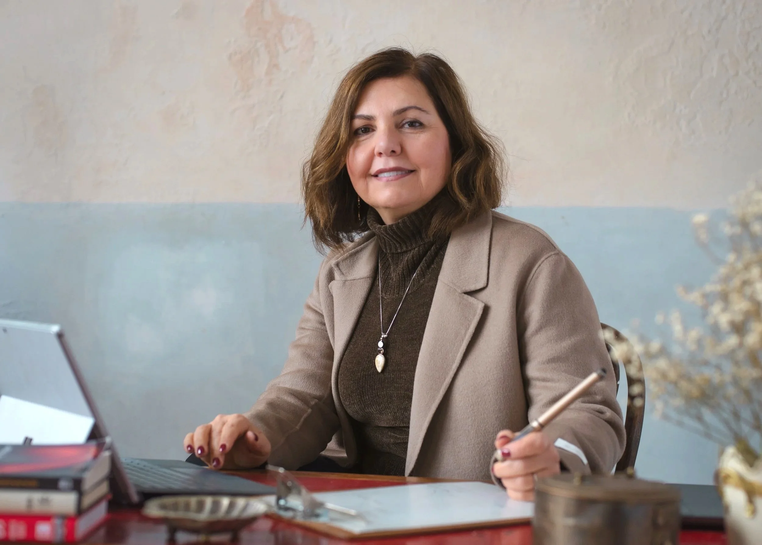 A woman with shoulder-length brown hair, wearing a tan blazer and dark turtleneck, sitting at a desk with a laptop, papers, and books, smiling at the camera.