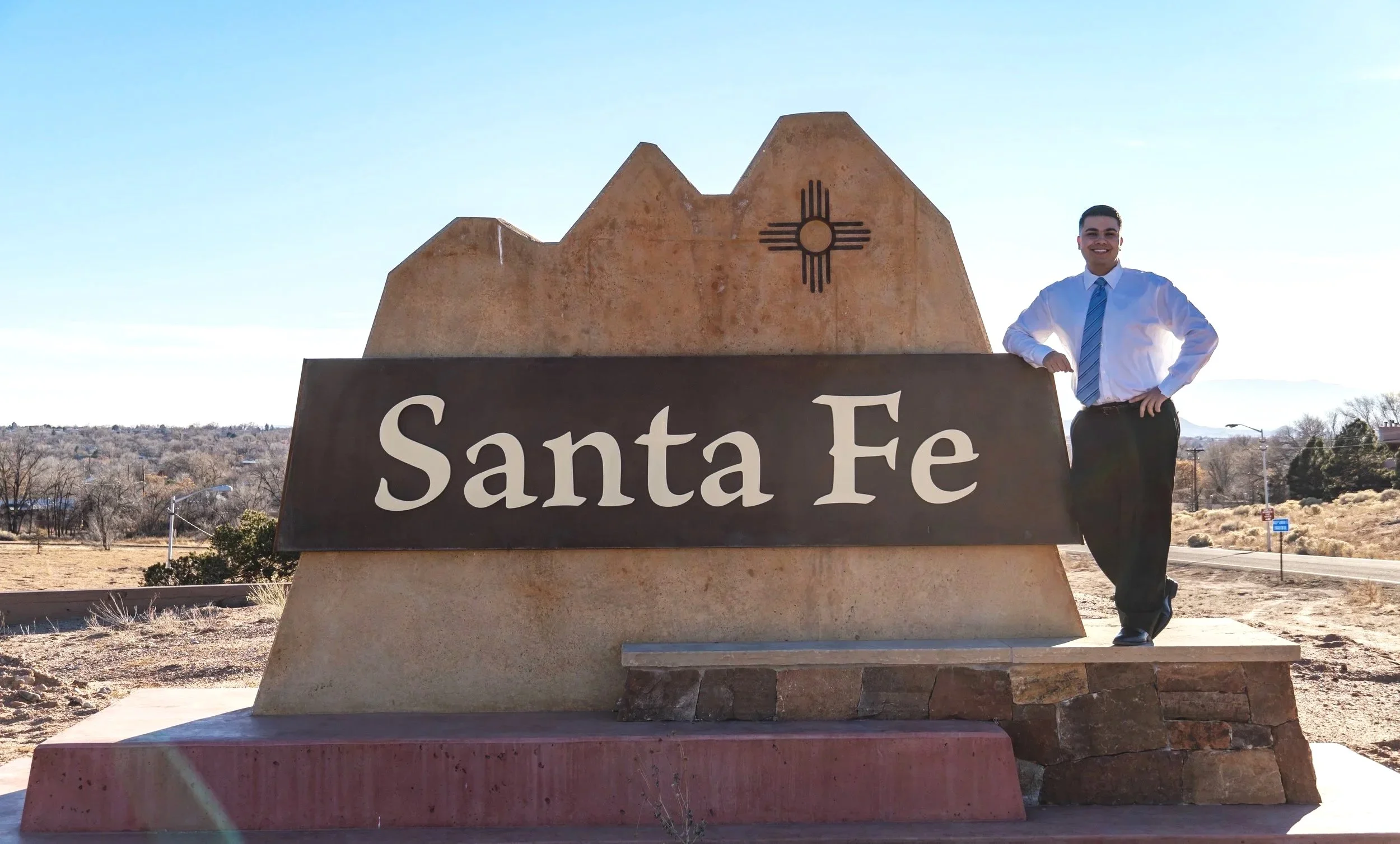 A man dressed in a white shirt and blue tie standing next to a large stone sign that says 'Santa Fe' in front of a desert landscape with trees and power lines.