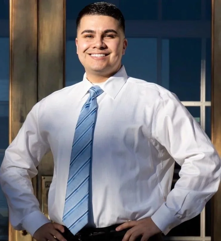 A man in a white dress shirt and blue striped tie standing indoors with hands on hips, smiling at the camera.