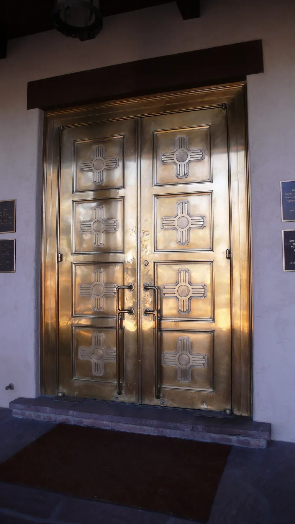 Large golden doors with a cross design on each panel, metal handles, and hardware, set within a church or religious building entrance.