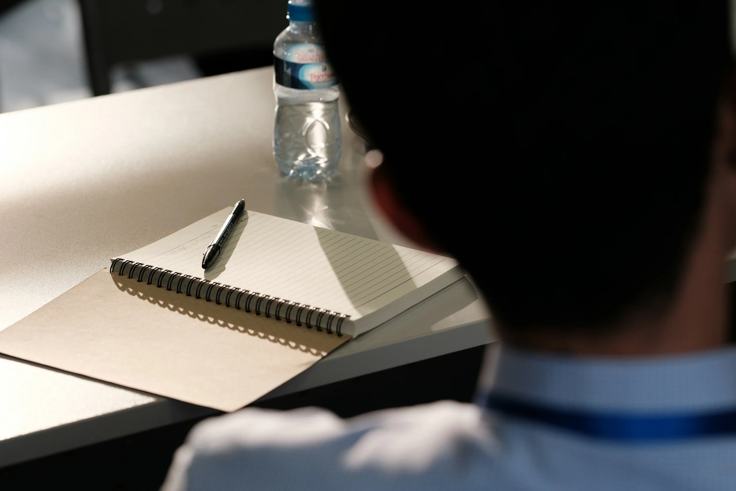 A notebook with lined pages and a pen resting on it, placed on a light-colored table. In the background, a partially visible water bottle and the edge of a chair are seen. The foreground shows the partial silhouette of a person's head.