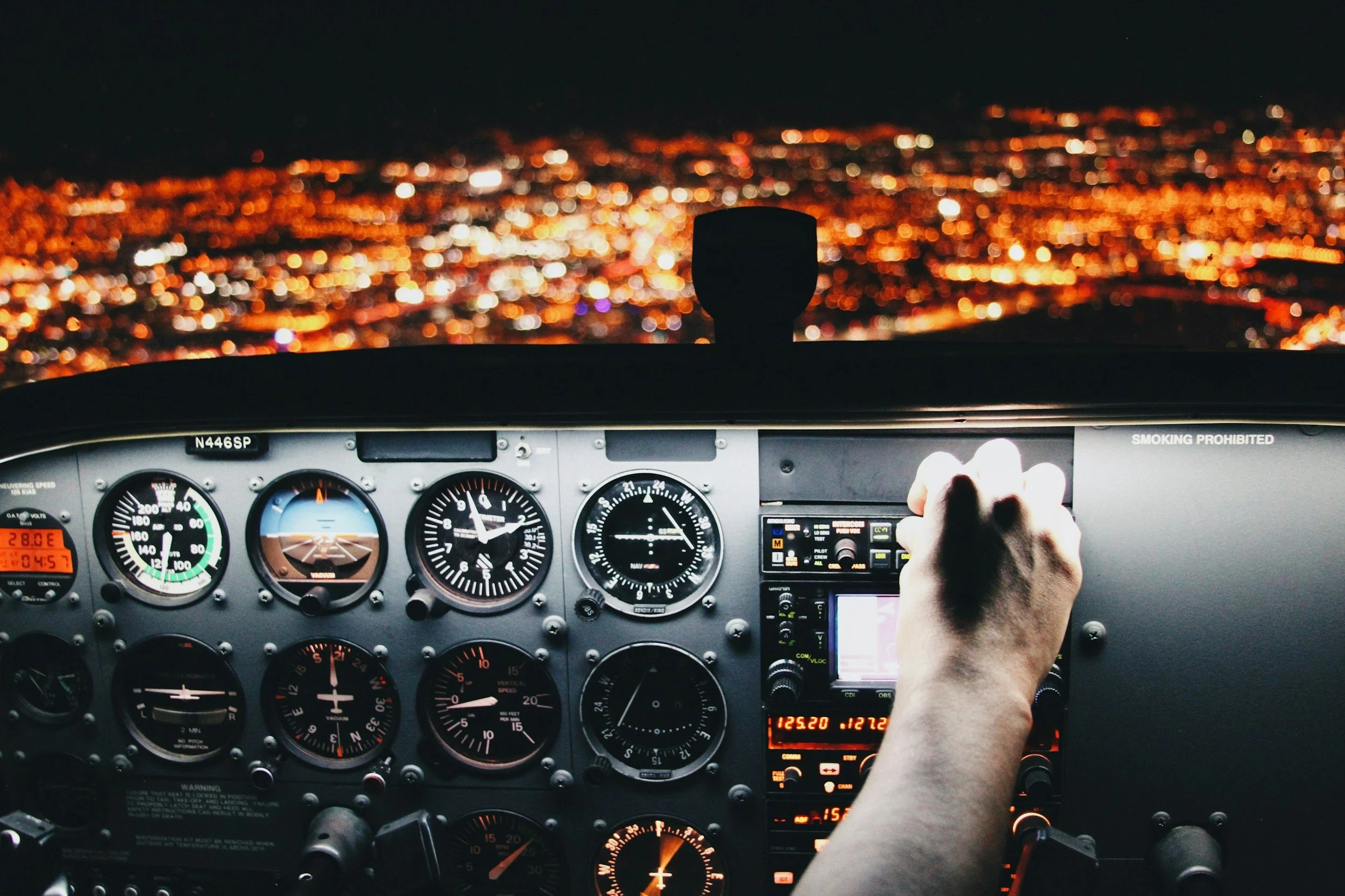 View from inside an airplane cockpit at night, showing the pilot's hand on the controls, with a horizon of city lights blurred in the distance.