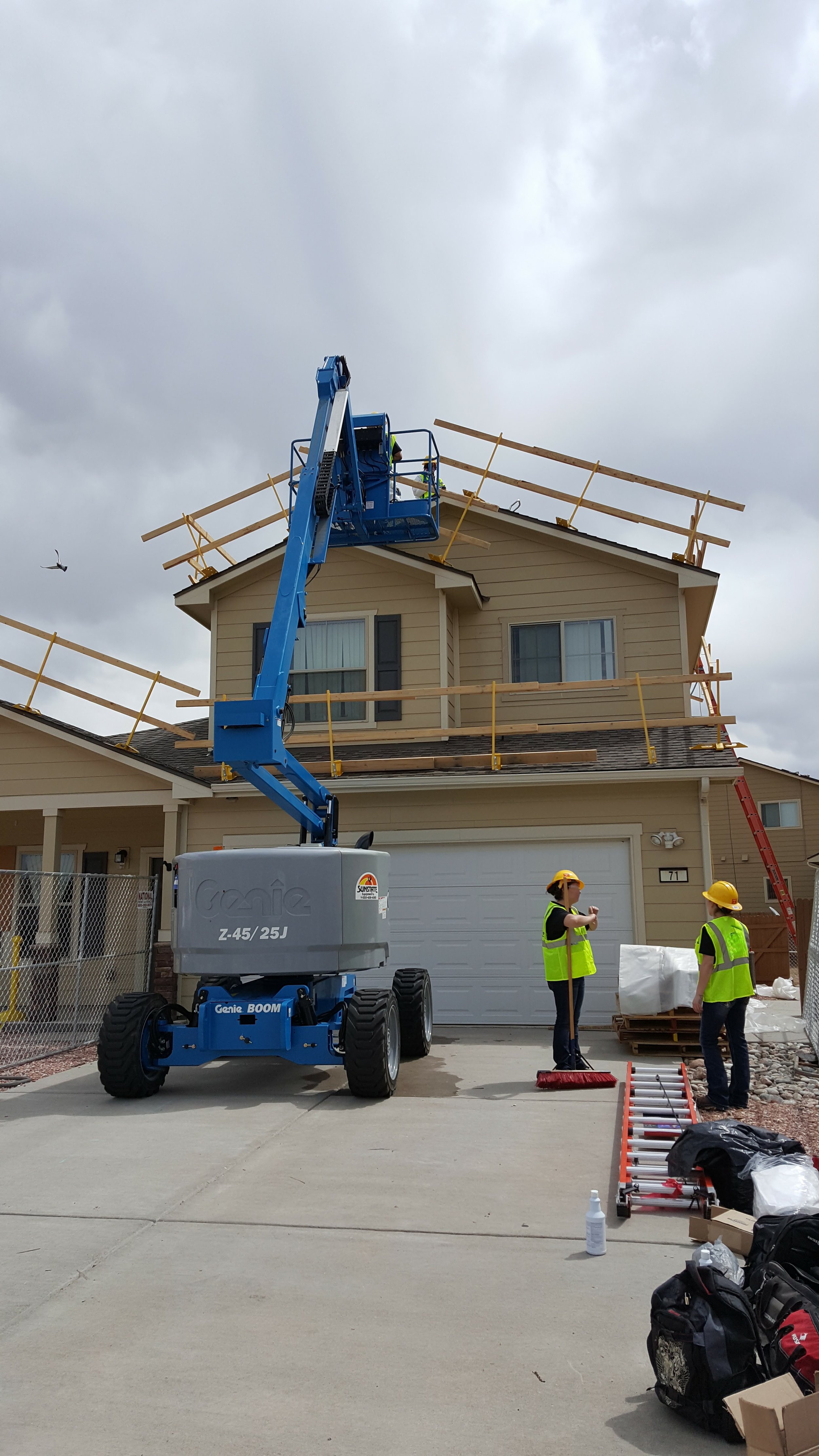 Construction workers using a lift on a residential house roof to install or repair the roof structure, with tools and equipment on the driveway in the foreground, and overcast sky above.