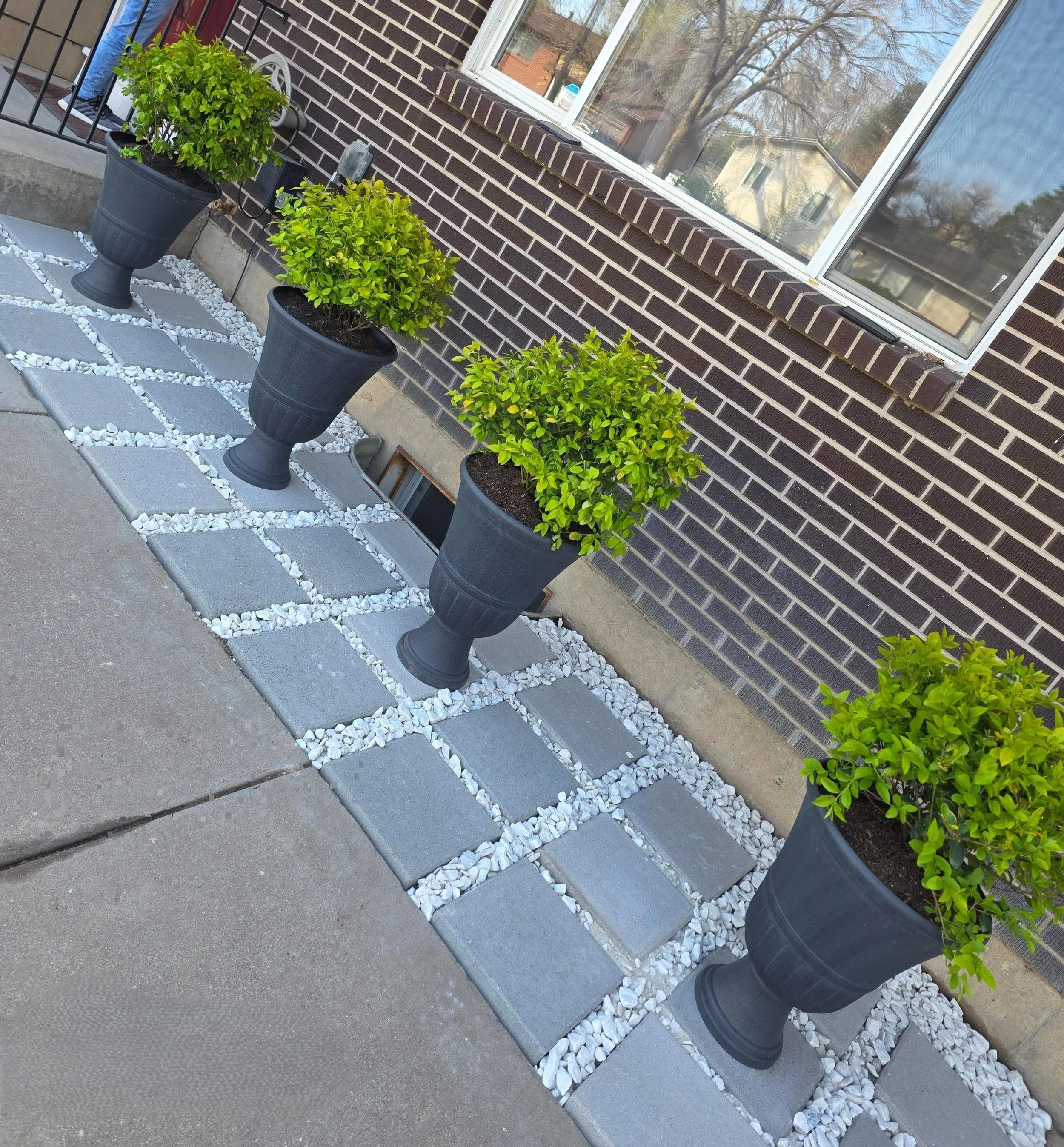A house exterior with three large planters with shrubs, a brick wall, a window, and a sidewalk with stone tiles and white gravel design.