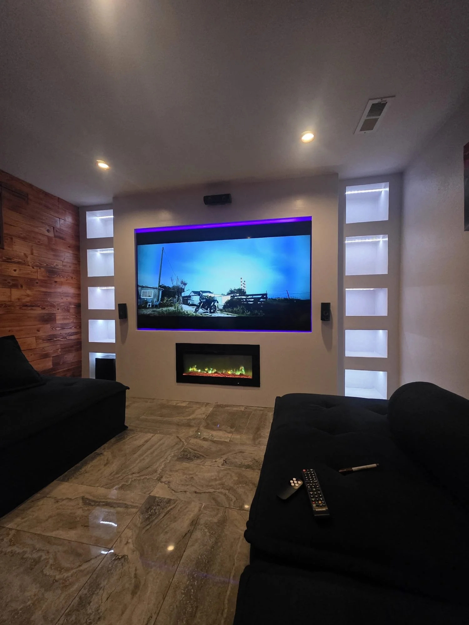 Living room with black sofas, a large flat-screen TV mounted on the wall displaying a scenery, LED strip lights around the TV, a fireplace below, and a wood-paneled wall on the left. The floor is tiled with beige and brown marbled tiles, and there is a remote and a TV remote on one of the sofas.