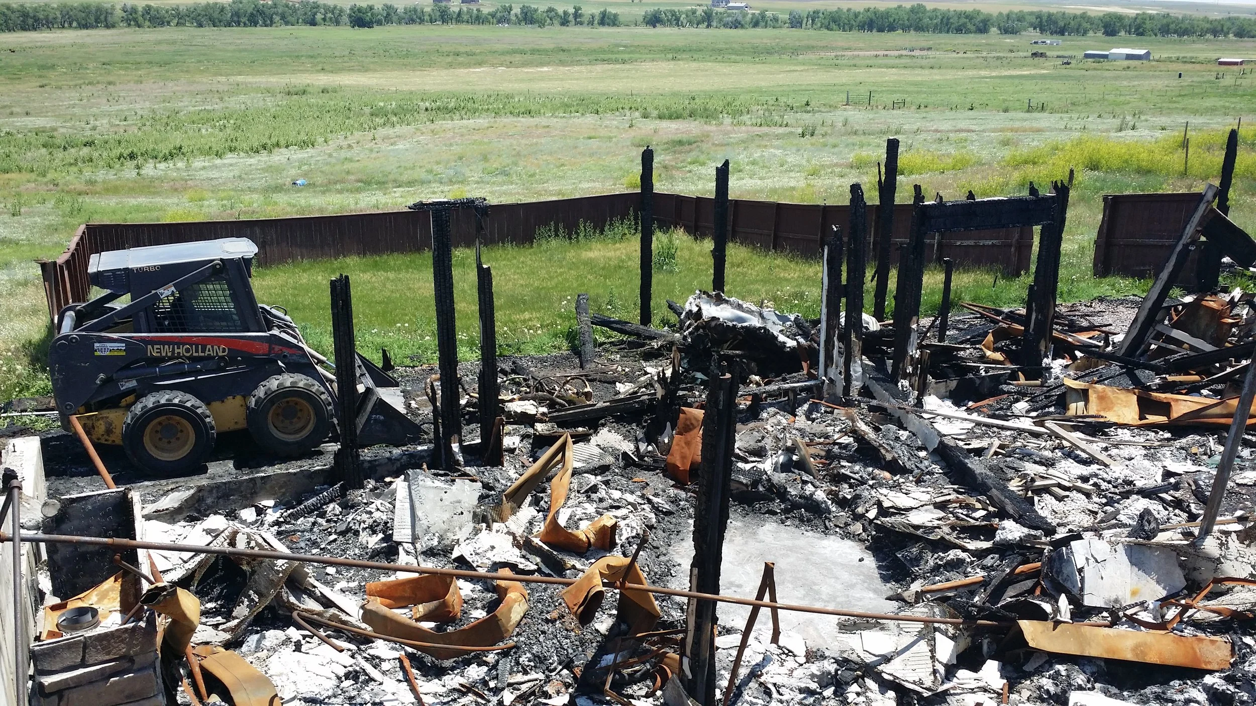 Burned remains of a building with charred wooden beams and rubble, a small construction vehicle on the left, enclosed by a fence, with green fields and a distant tree line in the background.