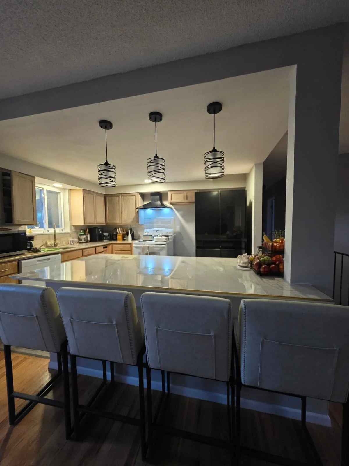 Kitchen with a white marble island counter, wooden cabinets, black appliances, a window, and three hanging pendant lights. Chairs are at the island, and fruit is on the counter.