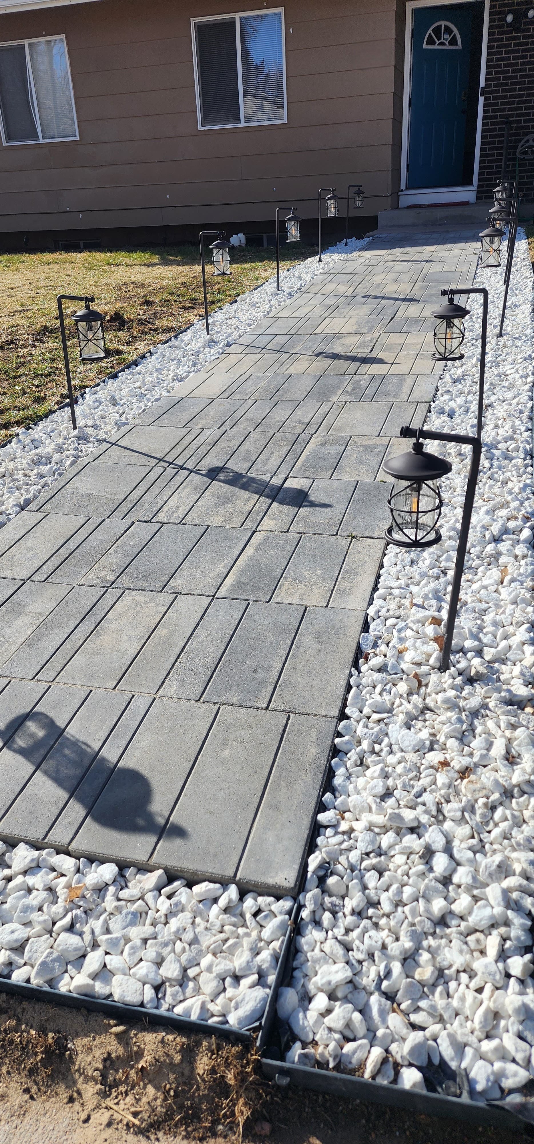 A newly paved concrete walkway with black metal lantern-style lights lining both sides, bordered by white decorative stones, leading up to a house with a blue front door and two windows.