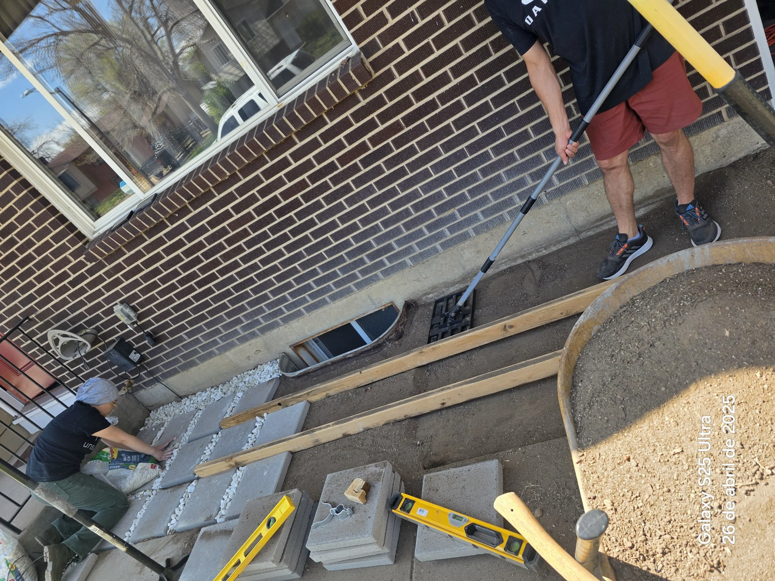 People working on building steps with concrete blocks outside a brick house, with tools and materials around.