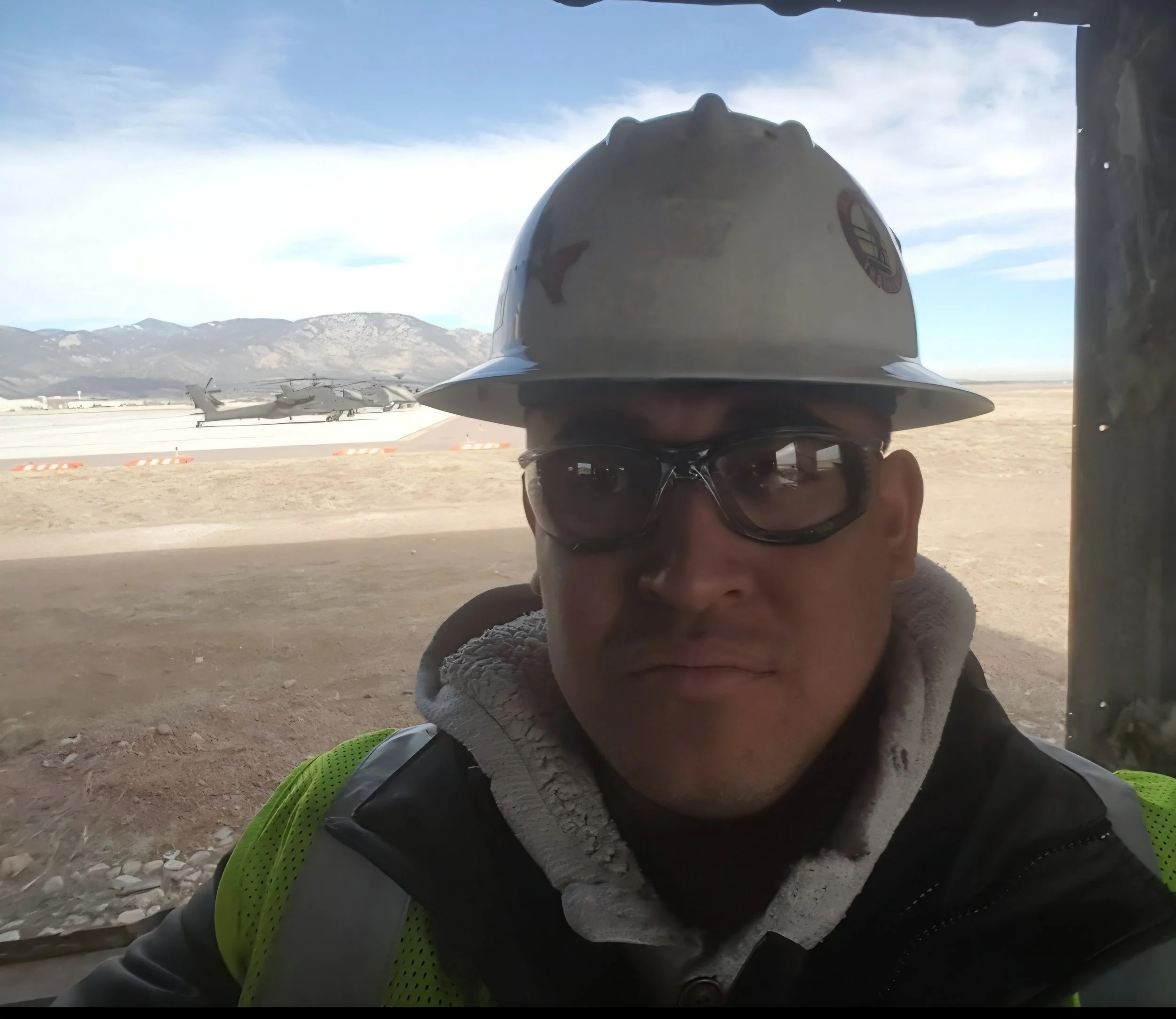 Person wearing sunglasses and a wide-brimmed hat, taking a selfie at an airport with airplane and mountain landscape in the background.