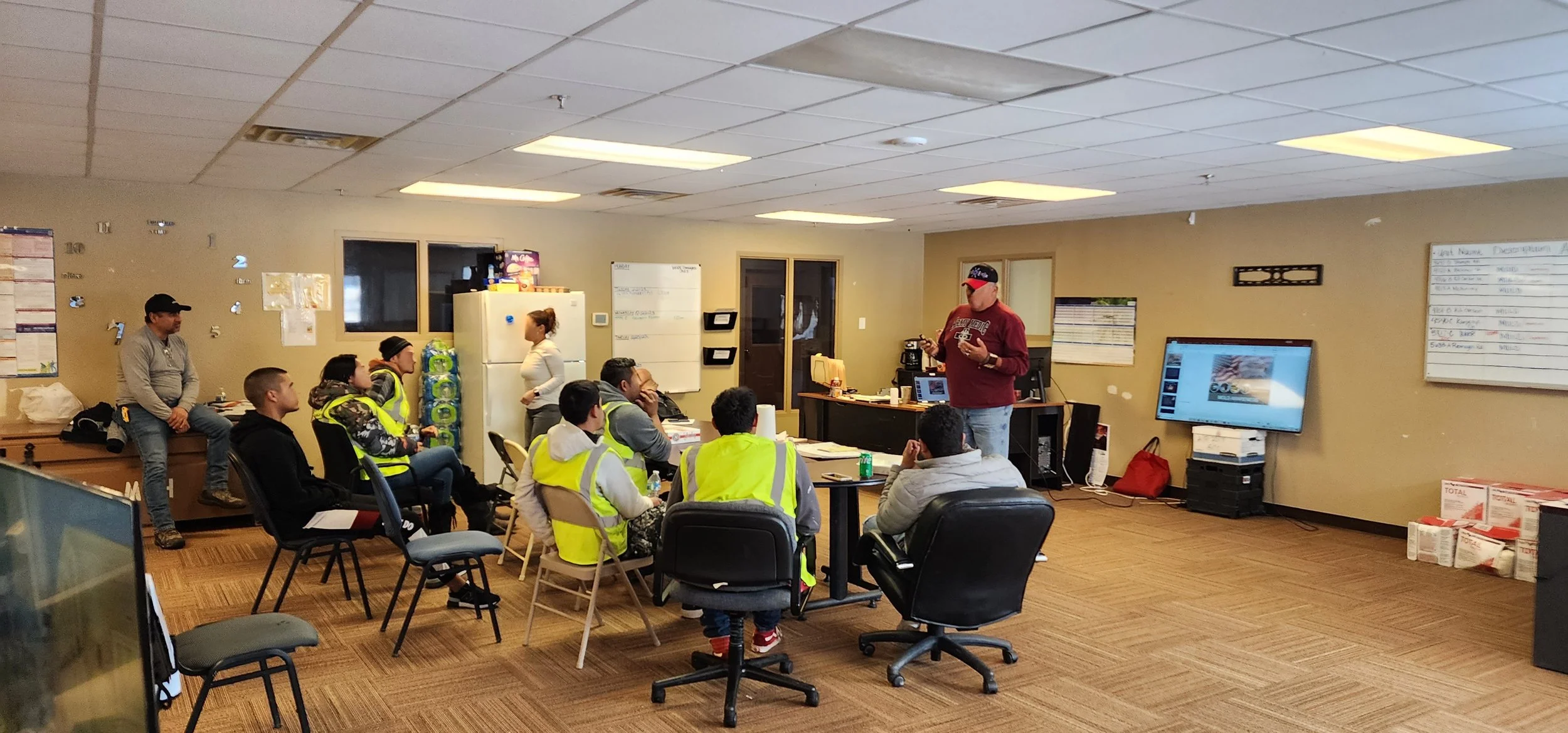 A classroom setting with a group of people wearing safety vests, seated and listening to a man in a red sweatshirt speaking and using a phone. Others are sitting or standing, and there is a whiteboard, a TV, and various office supplies in the background.