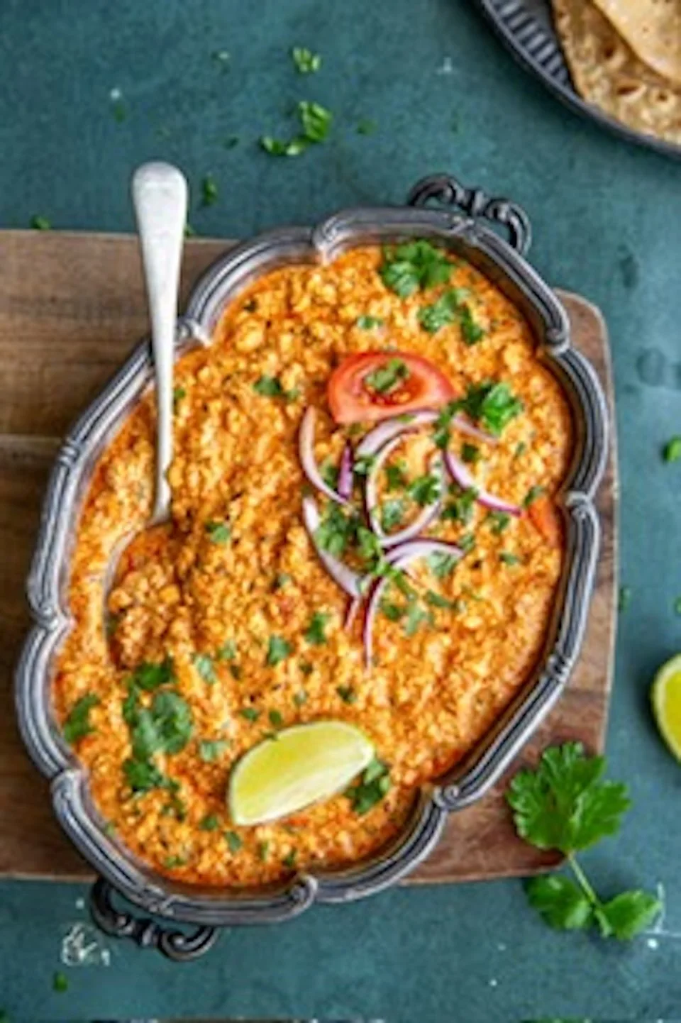 A serving dish with Mexican style shredded chicken in a bright orange sauce, garnished with cilantro, sliced red onions, a lime wedge, and a tomato slice. There is a spoon resting on the dish, and some cilantro on the surface nearby.