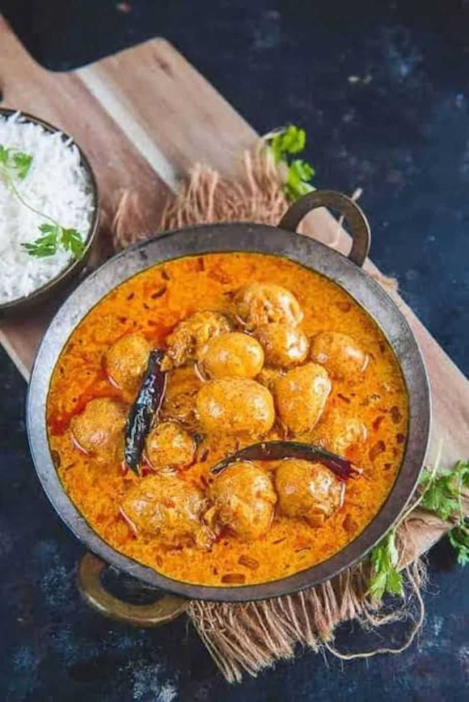 A bowl of Indian chicken curry with potatoes in a rich, orange-colored sauce, garnished with dried red chilies, served on a rustic tray with a side of white rice garnished with cilantro.