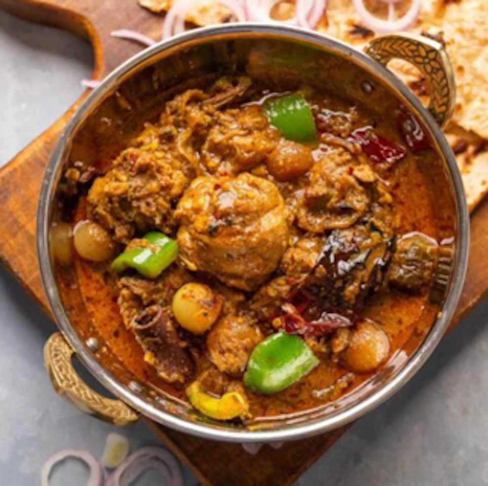 A bowl of Indian curry with meat, vegetables, and spices served on a wooden board, with naan bread and sliced onions on a grey surface.