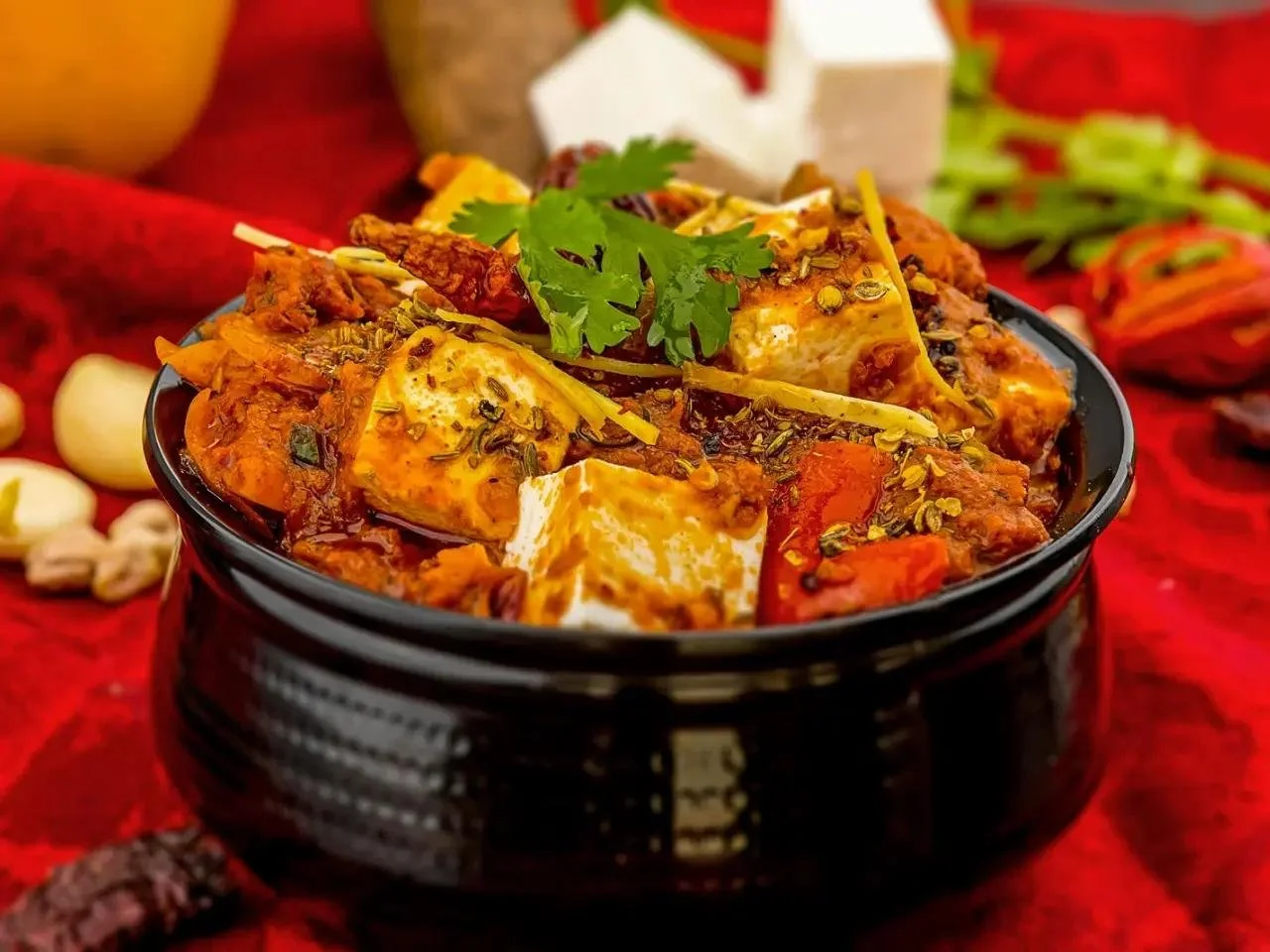 A bowl of Indian curry with tofu, garnished with cilantro, served on a red cloth, with garlic and spices around.