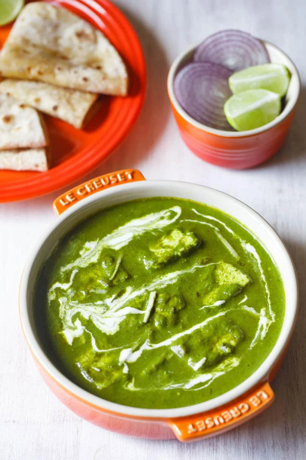 A bowl of green chicken enchilada sauce with shredded cheese, a cup of sliced red onion and lime wedges, and a basket of quesadillas.