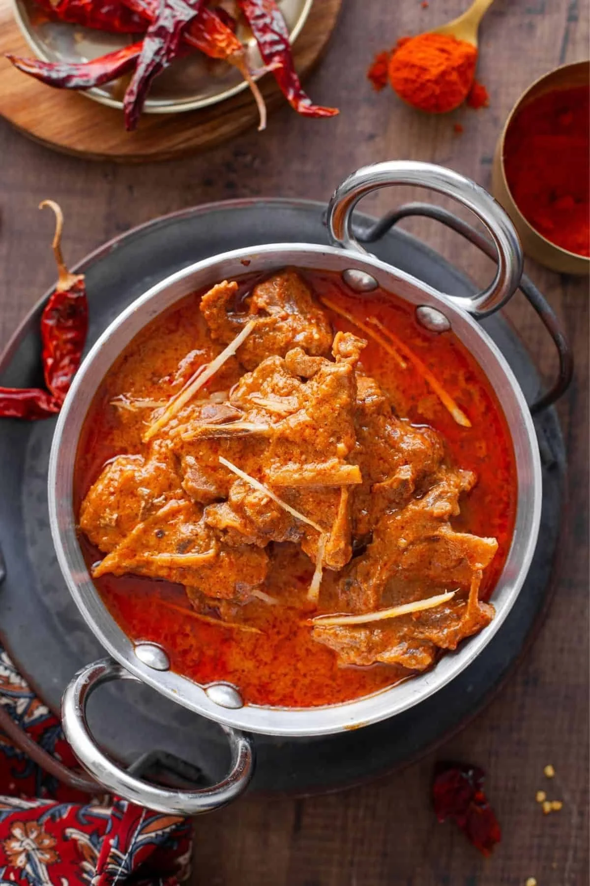 A stainless steel pot filled with a spicy curry containing meat and thin strips of ginger, sitting on a black tray. Surrounding the pot are dry red chilies, a small bowl of red chili powder, and a bowl of red sauce, with a wooden table underneath.