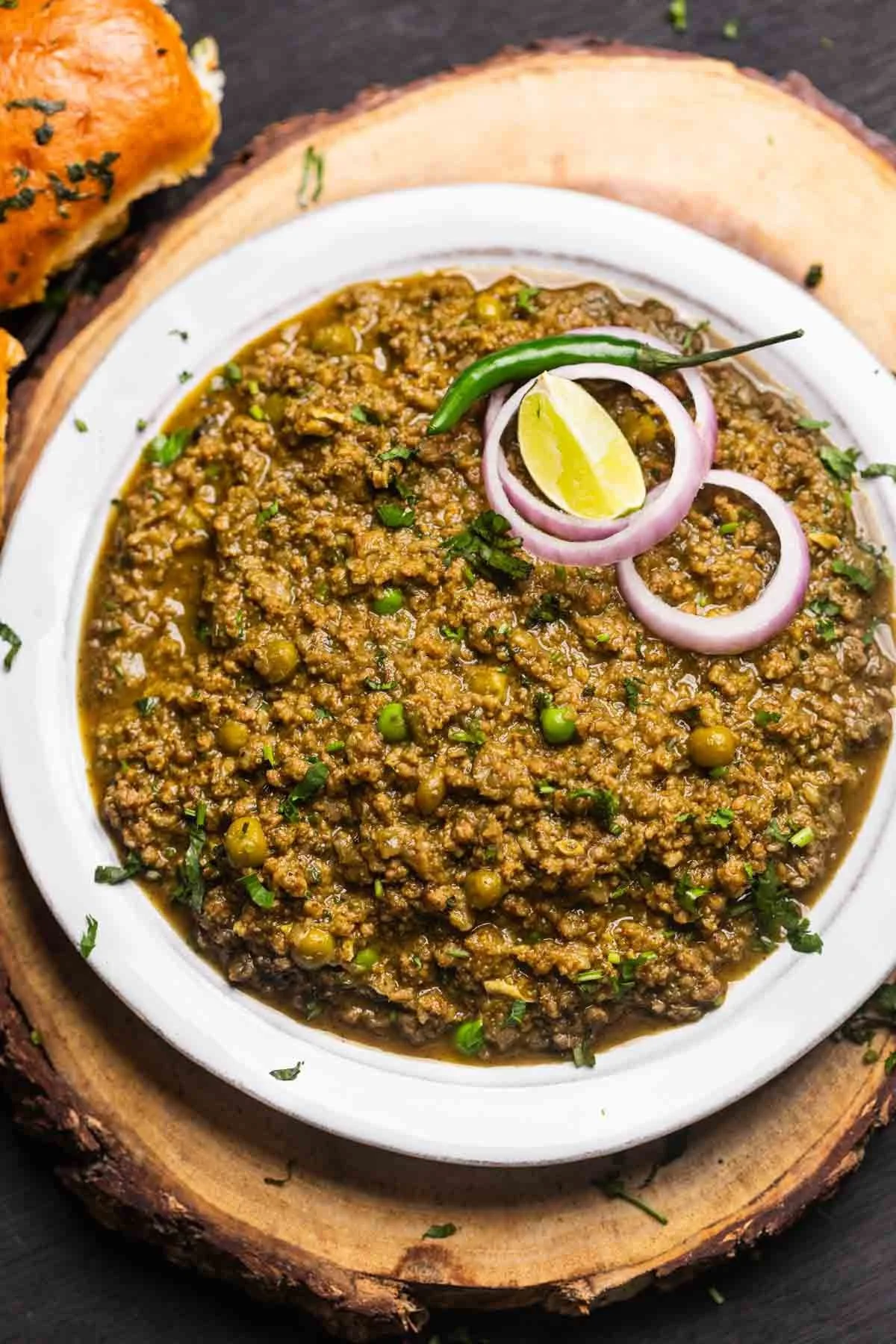 A bowl of minced meat curry garnished with sliced onions, a green chili, and a lemon wedge, served on a wooden platter with bread in the background.