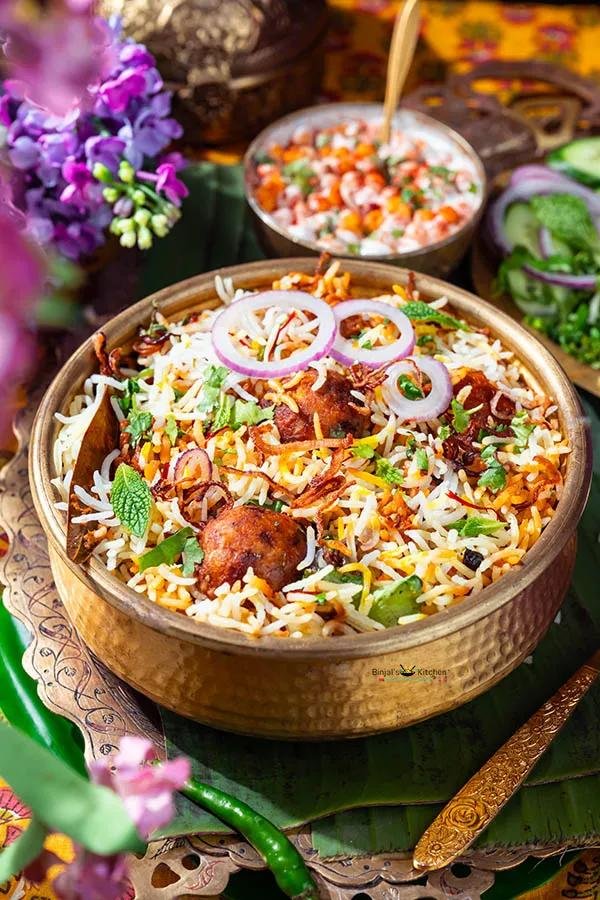 A bowl of Indian biryani topped with sliced onions and fresh herbs, garnished with fried onions, served on a plate with floral decorations and a small bowl of raita or curry in the background.