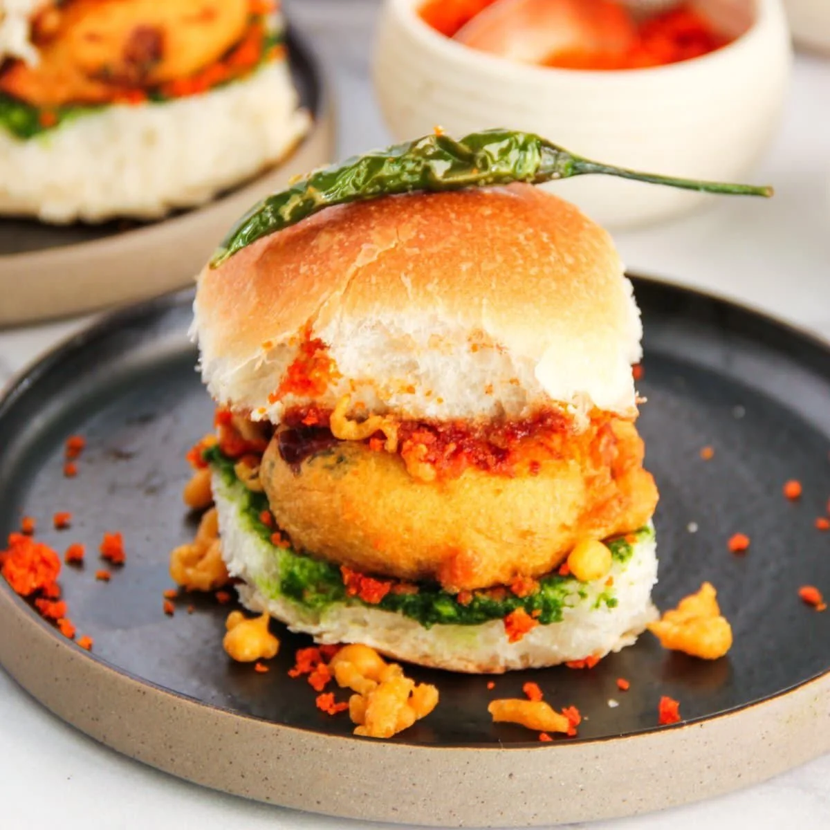Close-up of a bao bun sandwich with fried chicken, spicy sauce, green herbs, and a green chili on top, served on a black plate with scattered crumbs.