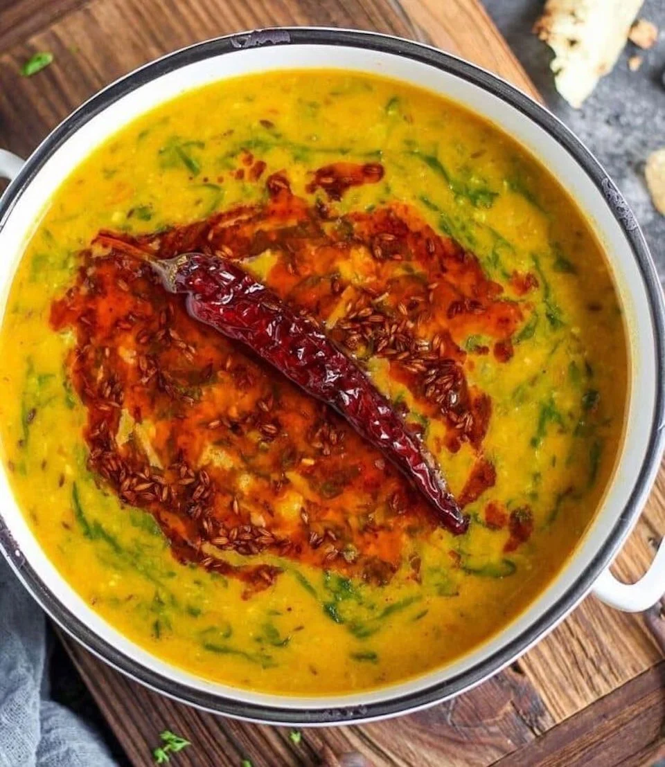 Close-up of a bowl of yellow lentil soup garnished with a dried red chili and red chili flakes on top, on a wooden surface.