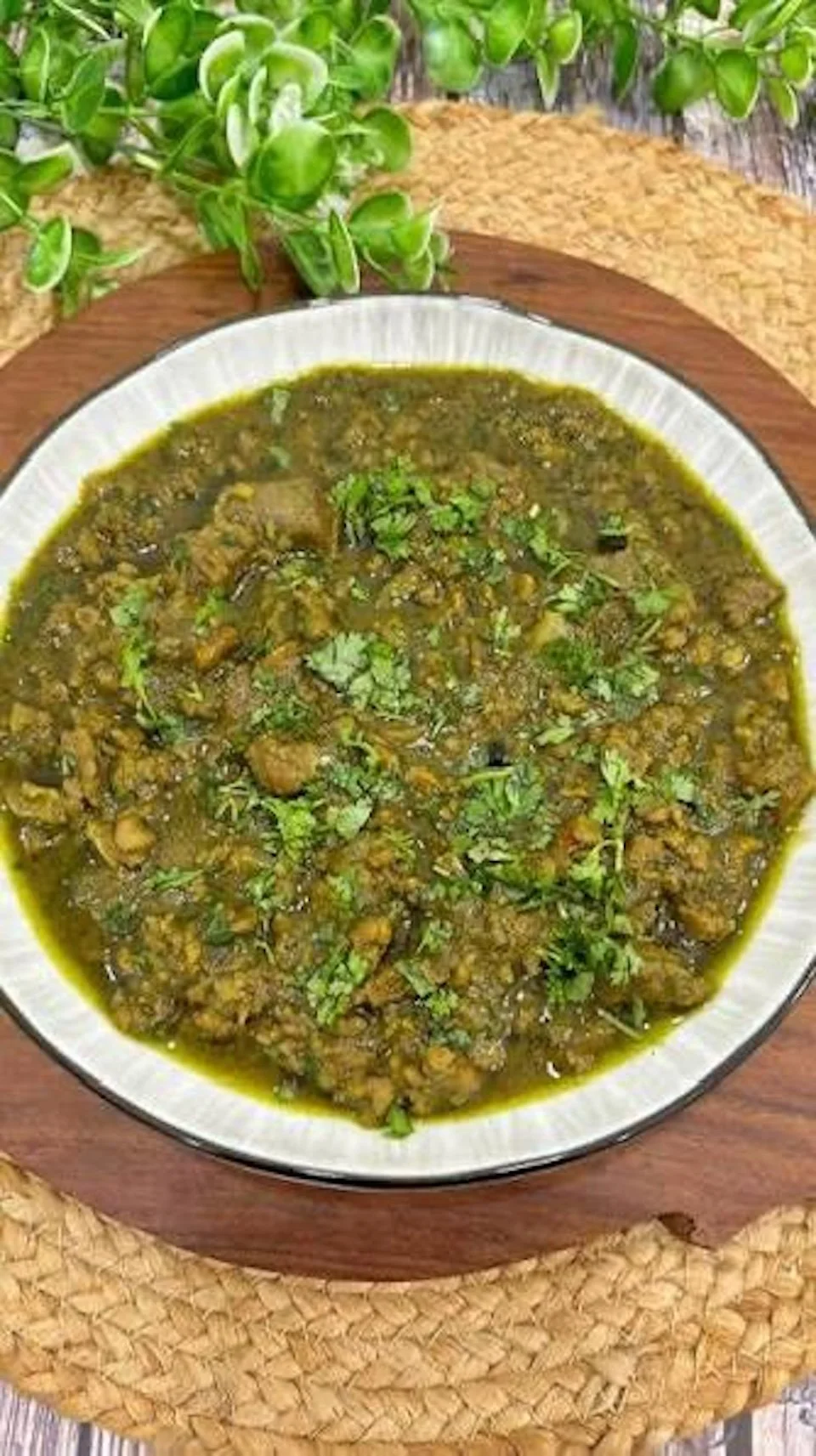 A bowl of cooked Indian-style lentil and vegetable curry garnished with chopped cilantro, placed on a wooden tray with greenery in the background.