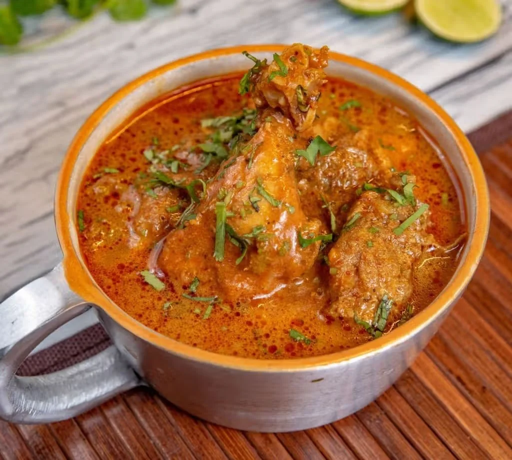 A bowl of Indian chicken curry garnished with chopped cilantro, set on a wooden table with lime wedges in the background.