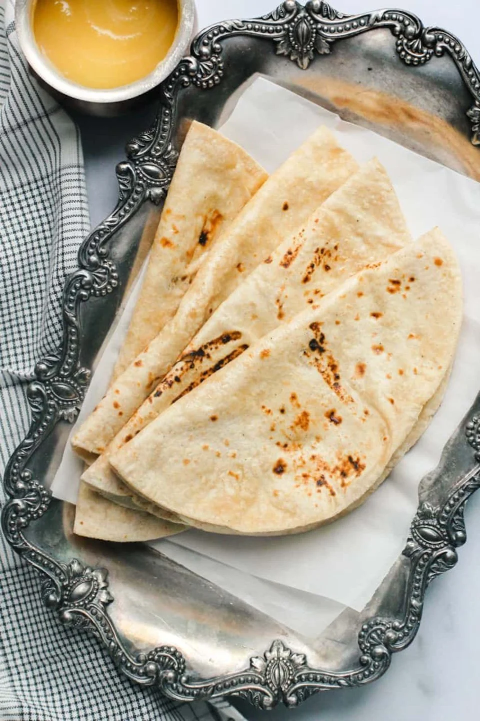 Three folded, toasted quesadillas on a decorative silver platter, with a small bowl of melted butter and a black and white checkered napkin.