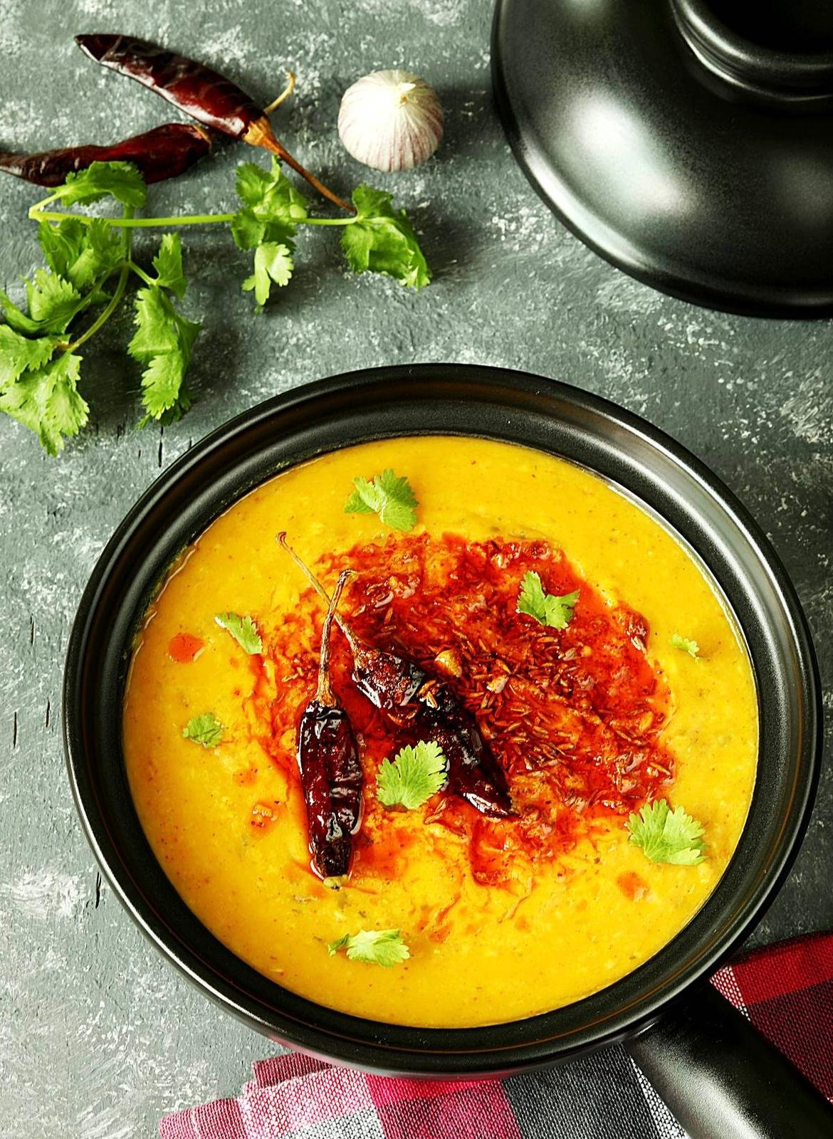 A bowl of yellow lentil soup topped with chili peppers, cilantro leaves, and a drizzle of red sauce. Surrounding ingredients include garlic, dried red chili peppers, fresh cilantro, and a black pot on a gray textured surface with a red and black chec