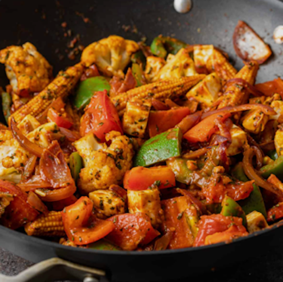 A close-up of a colorful vegetable stir-fry in a black skillet, including cauliflower, bell peppers, baby corn, onions, and tomatoes, seasoned and cooked.