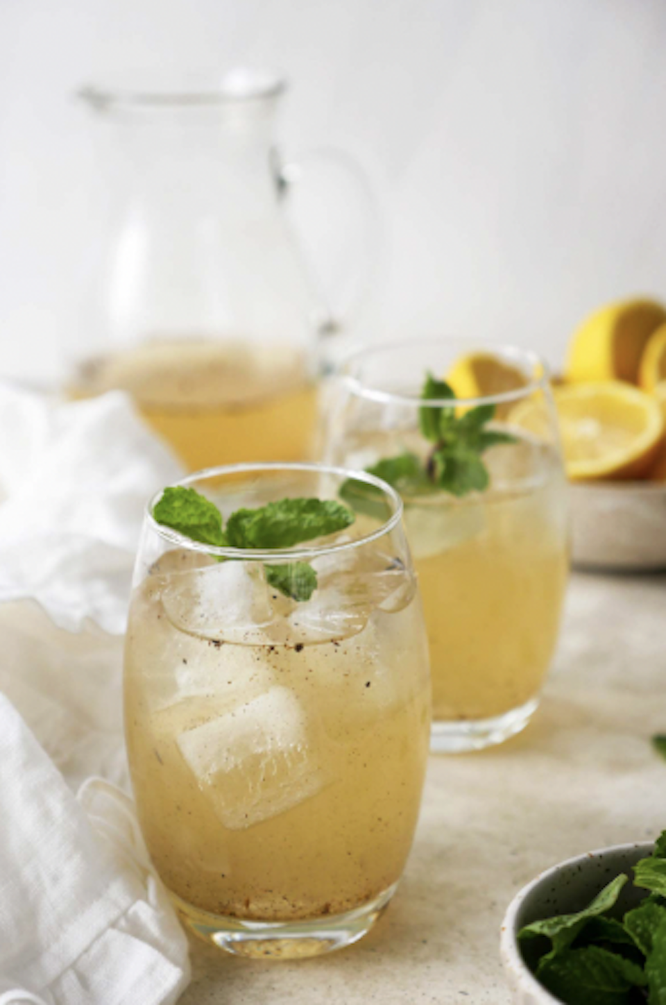 Two glasses of lemonade with ice and mint, a lemon half, and a pitcher of lemonade in the background on a white surface.