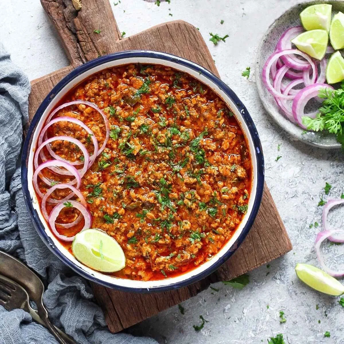 A bowl of chili garnished with chopped cilantro and sliced red onions, with lime wedges and additional sliced onions on a plate nearby.