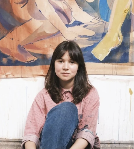 Young woman with long dark hair wearing a pink plaid shirt and jeans, sitting on a bench with a colorful mural in the background.