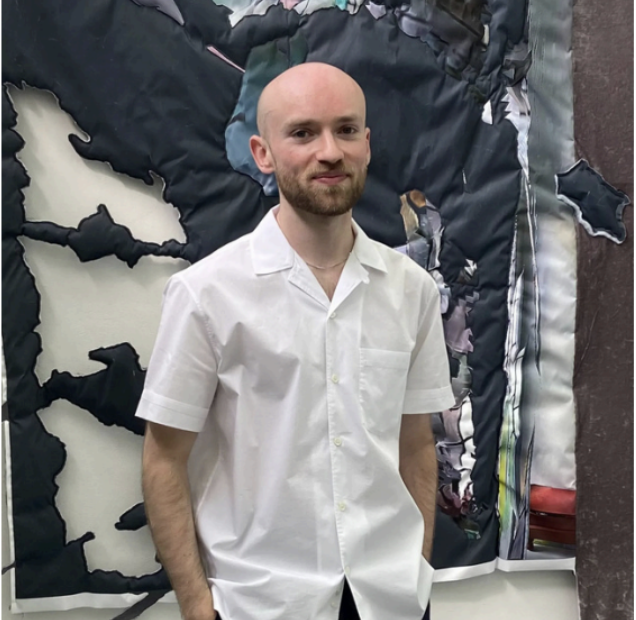 A young man with a shaved head and a beard, wearing a white short-sleeved shirt, standing indoors with a large abstract black and white artwork in the background.