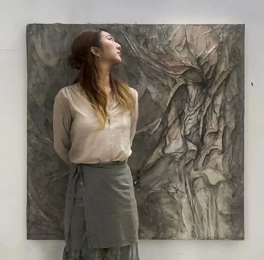 A woman with long hair, wearing a beige blouse and gray skirt, standing in front of an abstract, textured painting on a white gallery wall.