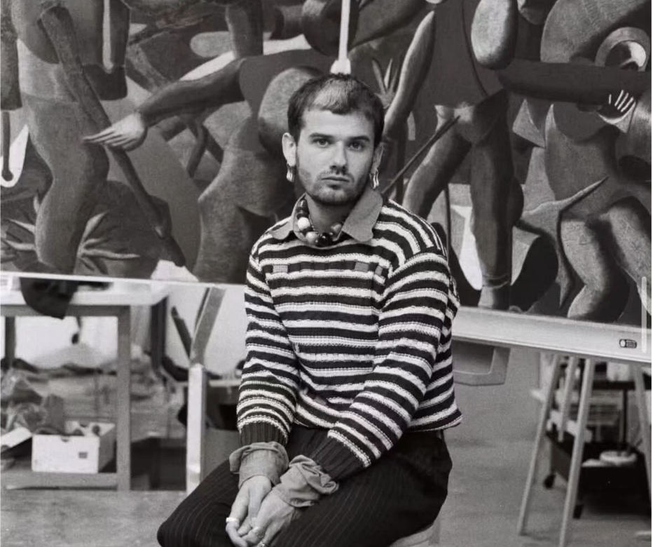 A young man with a beard, wearing a striped sweater and earrings, sitting with his hands clasped, in front of an abstract sculpture that resembles multiple human figures intertwined.