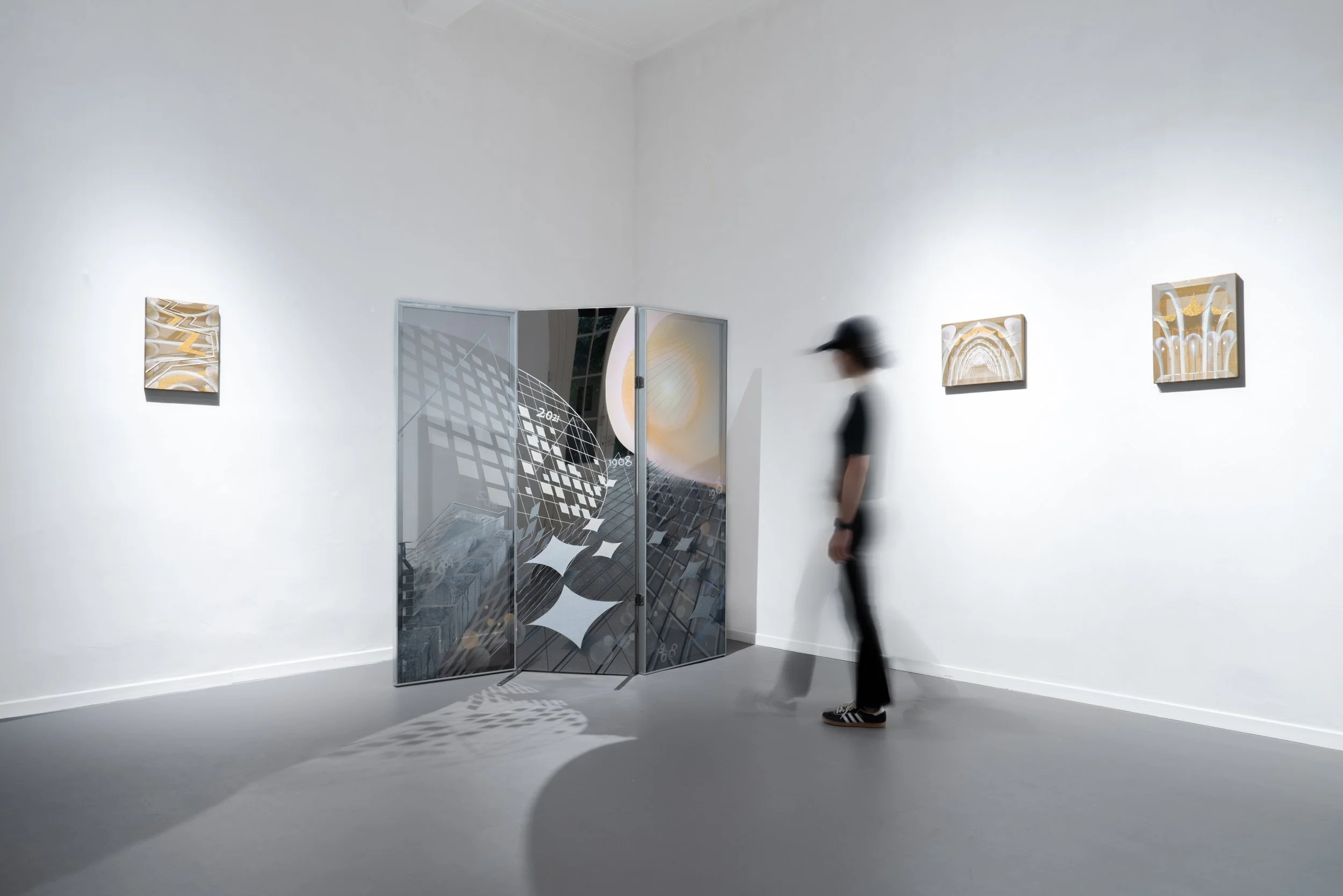 A person in black clothing and sneakers standing in an art gallery, observing artwork on the walls. The gallery has white walls and minimalistic decor, with artwork hung at eye level. The person appears to be slightly blurred, indicating movement.