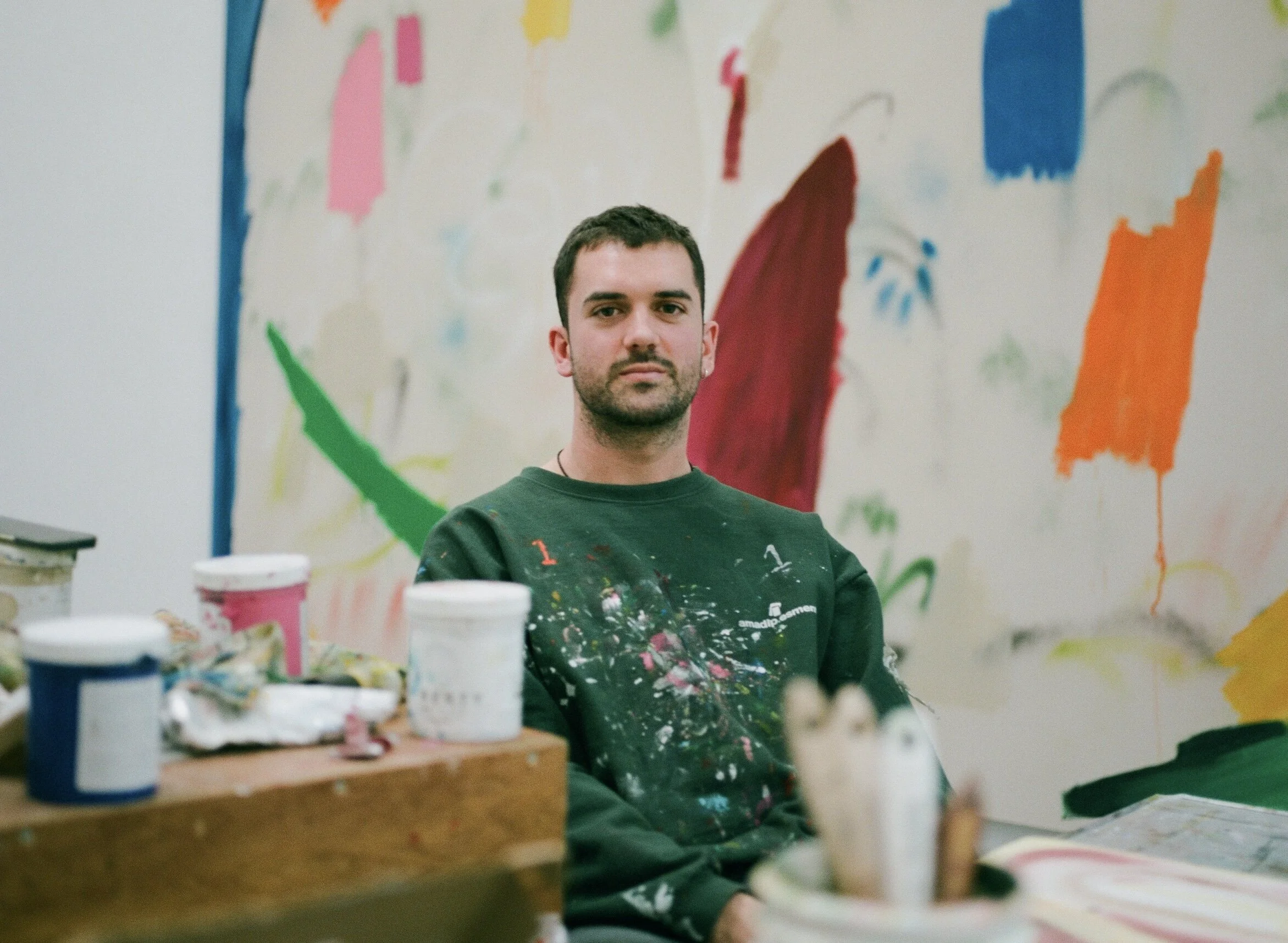 A man sitting in front of a colorful abstract mural. He is wearing a dark green shirt with paint splatters on it.