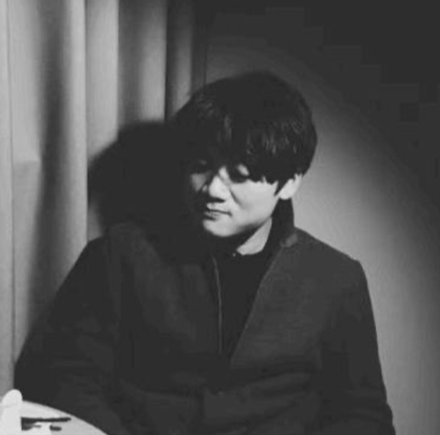 Black and white photo of a young man with dark hair, sitting near a wall and curtain, looking to the side.